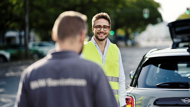 Vediamo un uomo con gilet giallo che sorride accanto a un'auto MINI, parlando con un tecnico BMW